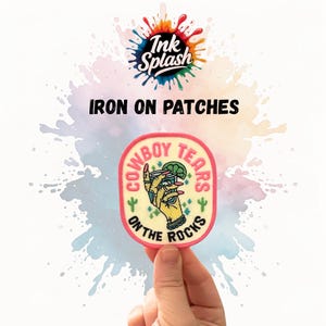 May include: An iron-on patch featuring the text "COWBOY TEARS ON THE ROCKS" and an illustration of a hand holding a drink. The patch has a light yellow background with a pink border and green cacti. The words "Ink Splash" are at the top.
