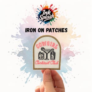 May include: An iron-on patch with a cream border and a pink and black design. The patch features the words "COWGIRL Cocktail Club" and cocktail illustrations. The patch is held by a hand, with the words "Ink Splash" and "IRON ON PATCHES" above.