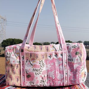 May include: A quilted tote bag with a floral pattern in shades of pink, white, and green. The bag has long, pink straps and a rectangular shape. The background is a field under a clear sky.