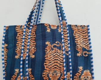 Könnte beinhalten: Eine große Tragetasche mit blauem Hintergrund und einem sich wiederholenden Muster aus orangefarbenen Tigern. Die Tasche hat blau-weiß gestreifte Griffe und Verzierungen. Die Tasche ist aus einem gesteppten Stoff gefertigt.