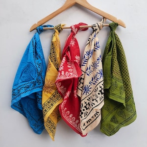 May include: Five patterned bandanas in blue, yellow, red, cream, and green hang from a wooden clothes hanger. Each bandana features a different black and white design. The bandanas are knotted at the top.