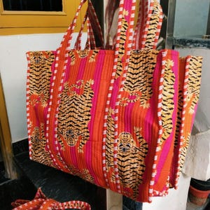 May include: Large tote bag with a vibrant tiger print. The bag features a quilted design with orange, pink, and white stripes. The tiger pattern is in black and yellow. The bag has two long handles.