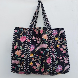 Op de afbeelding: Zwarte tote bag met een bloemenpatroon in roze, geel en paars. De tas heeft gestreepte handvatten en een gewatteerd ontwerp. De tas hangt tegen een witte achtergrond.