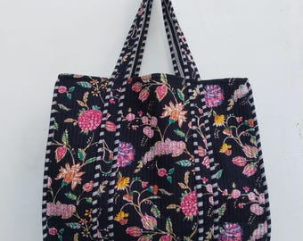 Könnte beinhalten: Schwarze Tragetasche mit einem Blumenmuster in Rosa, Gelb und Lila. Die Tasche hat gestreifte Griffe und ein gestepptes Design. Die Tasche hängt vor einem weißen Hintergrund.
