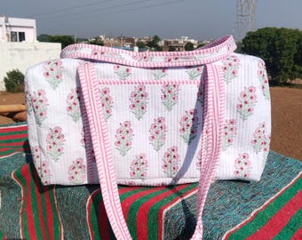 May include: White quilted tote bag with a floral pattern in pink and green. The bag has pink handles and trim. The bag is sitting on a striped textile. The bag is designed for carrying items.