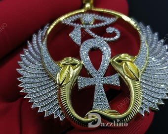 May include: A gold and silver pendant featuring an Ankh cross, Eye of Horus, and stylized wings. The pendant is encrusted with small, sparkling stones. The word "Dazzlino" is visible on the pendant.