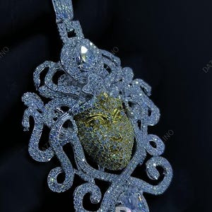 May include: A dazzling pendant featuring a Medusa design. The pendant showcases a gold-toned face with intricate detailing, surrounded by swirling, diamond-like embellishments. A large, clear gemstone is set at the bottom.