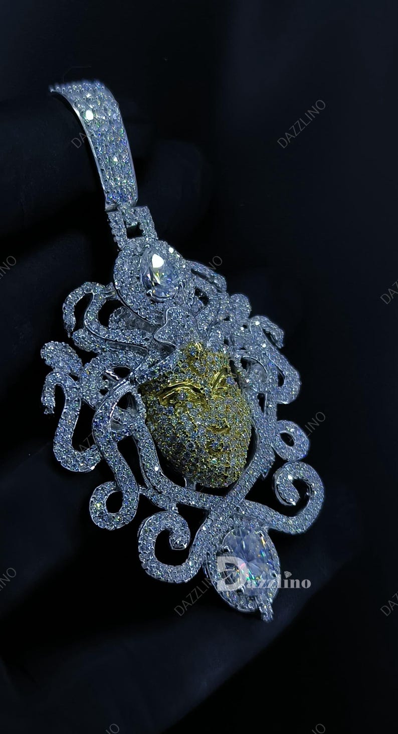May include: A dazzling pendant featuring a Medusa design. The pendant showcases a gold-toned face with intricate detailing, surrounded by swirling, diamond-like embellishments. A large, clear gemstone is set at the bottom.
