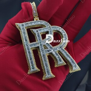 May include: A gold and silver pendant featuring the letters "FR" encrusted with small, rectangular-cut stones. The pendant is held against a red background. The word "Dazzlino" is visible.