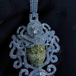 May include: A dazzling pendant featuring a detailed design with a central face-like element. The pendant is encrusted with numerous sparkling stones, with a mix of silver and gold tones. The design includes intricate, swirling patterns and a bail for a chain.