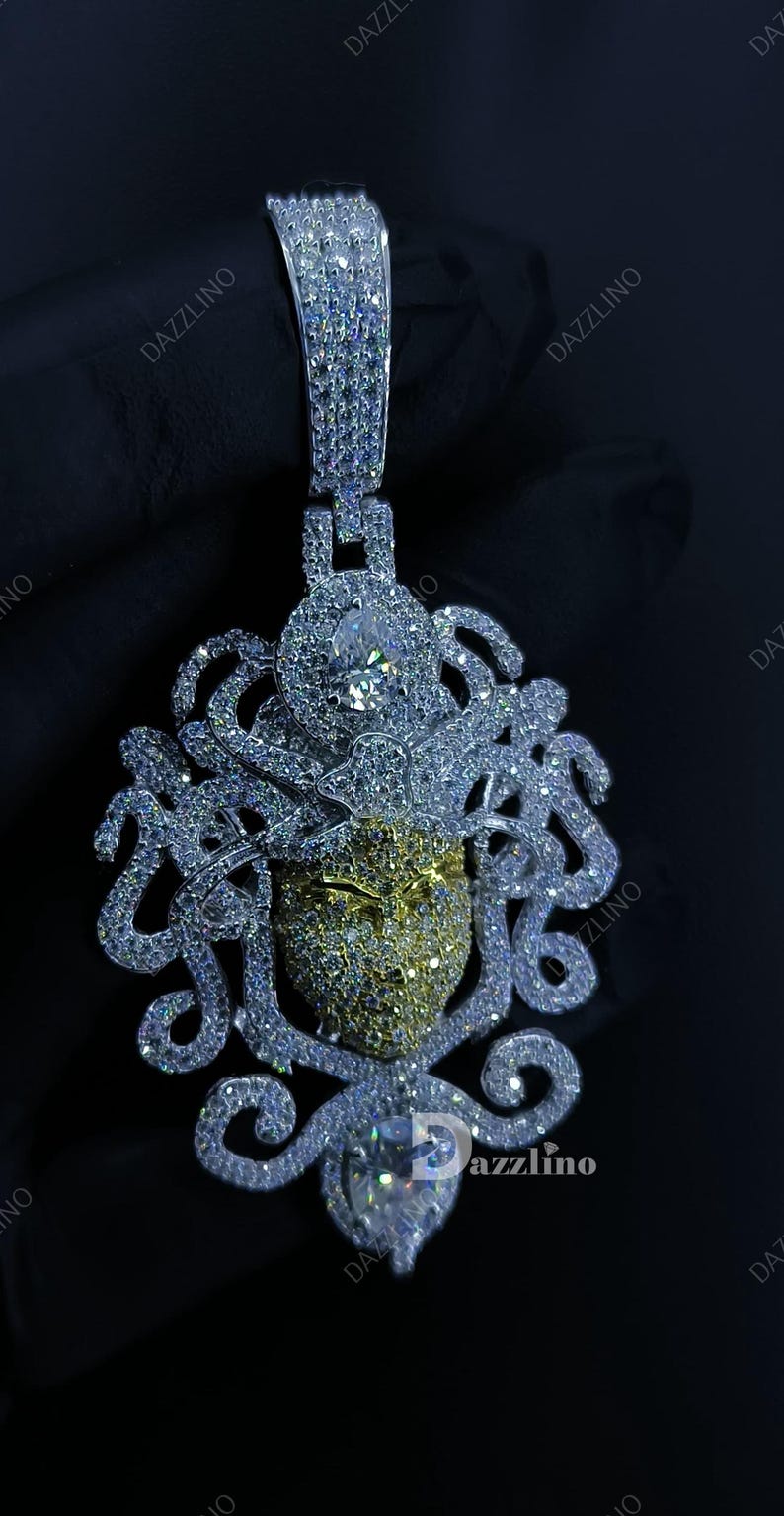 May include: A dazzling pendant featuring a detailed design with a central face-like element. The pendant is encrusted with numerous sparkling stones, with a mix of silver and gold tones. The design includes intricate, swirling patterns and a bail for a chain.