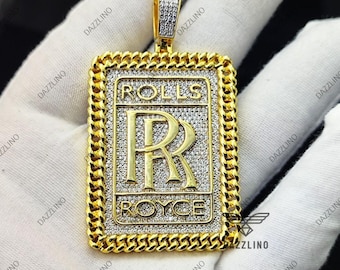 May include: A gold-colored rectangular pendant with a chain border. The pendant features the Rolls Royce logo with the text "ROLLS" above and "ROYCE" below the "RR" emblem. The pendant is encrusted with small, sparkling stones.