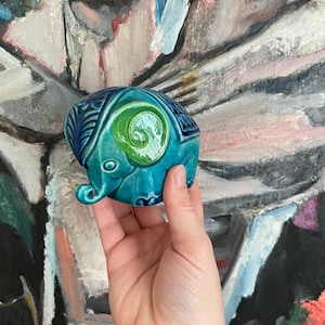 Peut inclure: Une petite figurine d'éléphant en céramique avec un glaçage bleu sarcelle et des accents verts. L'éléphant a un design détaillé sur son dos et une trompe enroulée.