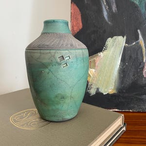 May include: A teal ceramic vase with a textured grey rim and a small square design. The vase is placed on a book with gold lettering, with an abstract painting in the background. The vase has a unique, organic shape and is a decorative item.