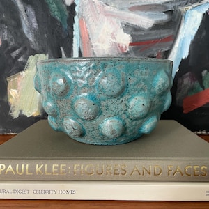 May include: A teal blue ceramic planter with a textured surface and a pattern of raised circles. The planter is sitting on top of two books. The top book has the title "Paul Klee: Figures and Faces" in gold lettering.