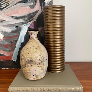 May include: Two vases, one a speckled brown and white ceramic vase and the other a tall, gold, ribbed metal vase, are displayed on a wooden surface in front of an abstract painting. The vases are sitting on top of two books, one with the title "Paul Klee: Figures and Faces" and the other with the title "Klee".
