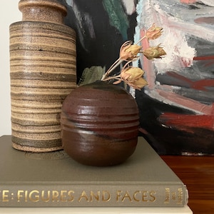 May include: Two ceramic vases, one brown and one with brown and beige stripes, are on a grey book with the title "L Klee: Figures and Faces" in gold lettering. The brown vase has dried flowers inside.