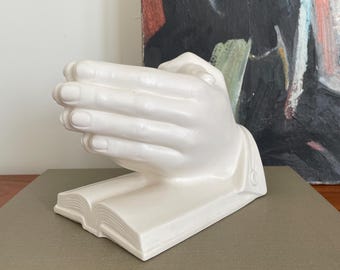 May include: White ceramic sculpture of two hands clasped together in prayer, resting on a closed book. The sculpture is on a brown surface.