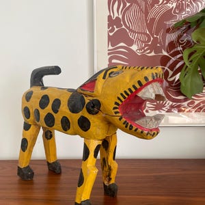 May include: A hand-carved wooden jaguar figurine. The jaguar is painted yellow with black spots and has a black tail. The mouth is open, revealing white teeth and a red interior. The figurine is approximately 25 cm long.