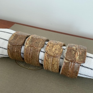 May include: Four ceramic napkin rings in a warm brown colour, each with a textured, speckled surface. The rings are arranged around a striped napkin, suggesting a rustic or farmhouse style for table settings.