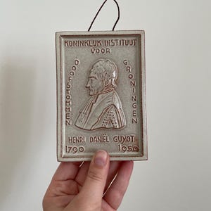 May include: An off-white ceramic plaque featuring a portrait of Henri Daniel Guyot. The plaque has embossed Dutch text, including the years 1790 and 1950. A brown cord is attached for hanging.
