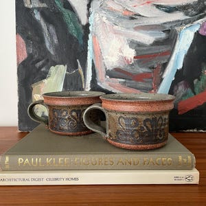 May include: Two ceramic mugs with handles, in brown and tan tones, adorned with a floral pattern. They rest on a book titled "PAUL KLEE: FIGURES AND FACES". An abstract painting provides the backdrop to the scene.