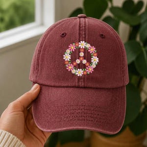 May include: A burgundy baseball cap with a peace sign embroidered on the front. The peace sign is made of colorful flowers in shades of pink, white, and yellow. The cap has a curved brim and an adjustable strap.