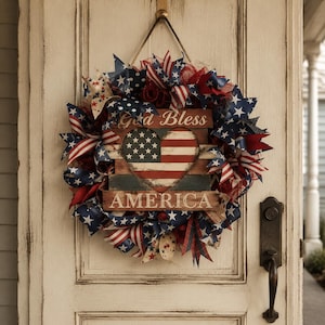 May include: A patriotic wreath with red, white, and blue ribbons and star accents. The wreath features a wooden heart with the American flag design and the words "God Bless AMERICA". It hangs on a white door.