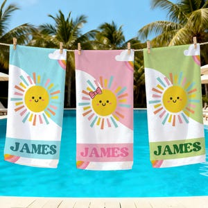 May include: Three personalized beach towels hanging on a clothesline. Each towel features a smiling sun design with colorful rays, a rainbow, and the name "JAMES" in a bold font. The towels are in shades of blue, pink, and green.