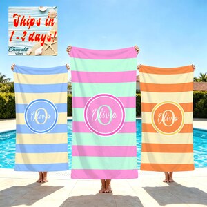 May include: Three rectangular beach towels featuring horizontal stripes and a central circular monogram. The left towel is blue and cream, the center is mint green and pink, and the right is orange and cream. Each displays the name Olivia.