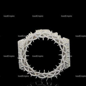 May include: A silver ring featuring a crown of thorns design, studded with small, glittering stones. The thorns are sharply defined. The ring is set against a black backdrop, with the brand name "IcedEmpire" repeated around the edges.