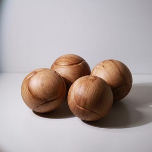 Op de afbeelding: Vier houten bollen met een lichtbruine afwerking zijn gerangschikt op een wit oppervlak. Elke bol heeft een gesneden ontwerp en een natuurlijke houtnerf. De objecten worden verlicht met een schijnwerper, waardoor schaduwen ontstaan.