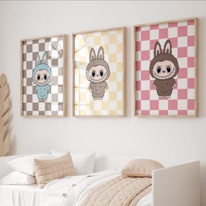 May include: Three framed prints featuring cartoon characters in bunny suits on checkered backgrounds. The prints are in shades of blue, brown, and pink. The frames are made of light-colored wood, and the prints are displayed above a bed.