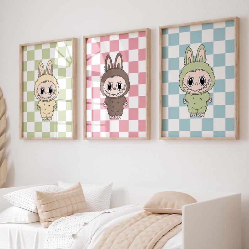 May include: Three framed art prints featuring cartoon characters with bunny ears and a toothy grin. Each character is set against a checkered background in green, pink, and blue. The prints are displayed above a bed with pillows and a blanket.