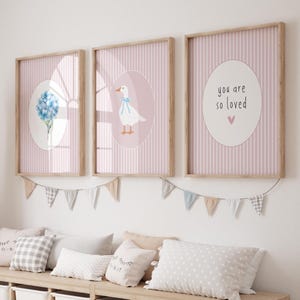 Puede incluir: Tres impresiones enmarcadas con un fondo a rayas rosa claro. Una impresión presenta una hortensia azul, otra un ganso blanco con un lazo azul, y la tercera dice "you are so loved" con un corazón rosa. Una guirnalda de pequeñas banderas cuelga debajo.