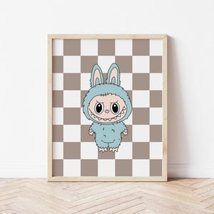 May include: Framed artwork featuring a cartoon monster in a blue bunny costume. The monster has a wide, toothy grin and is set against a brown and white checkered background. The artwork is in a light wooden frame.