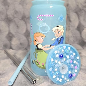 May include: A light blue tumbler featuring a cartoon image of two characters. The tumbler has a matching lid with decorative elements and comes with a reusable straw and cleaning brush. The design includes snowflakes and candy cane graphics.