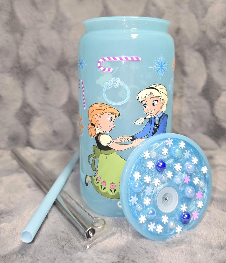 May include: A light blue tumbler featuring a cartoon image of two characters. The tumbler has a matching lid with decorative elements and comes with a reusable straw and cleaning brush. The design includes snowflakes and candy cane graphics.