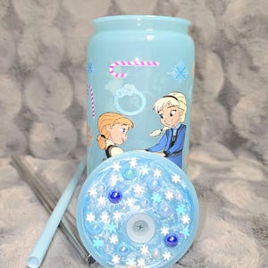 May include: A light blue tumbler with a matching lid and straw. The tumbler features a design of two cartoon characters, snowflakes, and a candy cane. The lid is decorated with blue and white embellishments.