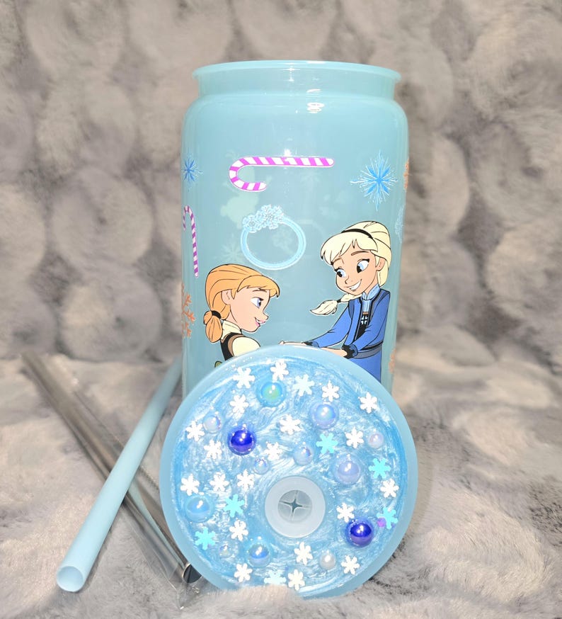 May include: A light blue tumbler with a matching lid and straw. The tumbler features a design of two cartoon characters, snowflakes, and a candy cane. The lid is decorated with blue and white embellishments.