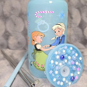 May include: A light blue tumbler featuring illustrations of two animated characters, candy canes, snowflakes, and rings. The tumbler includes a matching lid with decorative elements and a straw. A straw cleaner is also included.