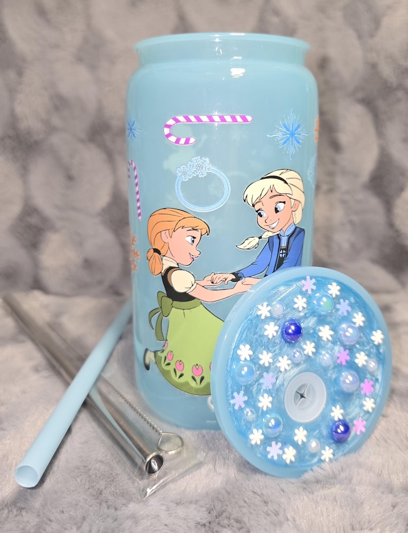 May include: A light blue tumbler featuring illustrations of two animated characters, candy canes, snowflakes, and rings. The tumbler includes a matching lid with decorative elements and a straw. A straw cleaner is also included.