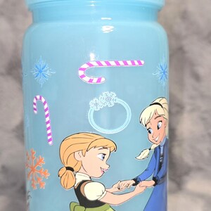 May include: A light blue tumbler with a matching straw, featuring illustrations of two animated characters. The tumbler is decorated with snowflake and candy cane stickers. The characters are wearing dresses.