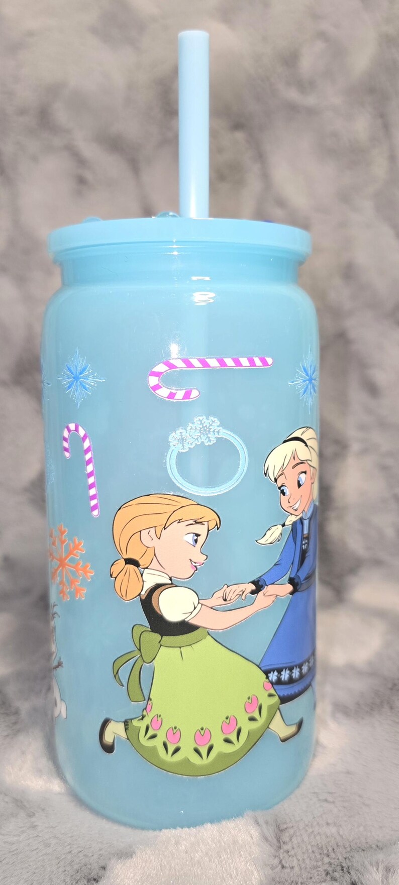 May include: A light blue tumbler with a matching straw, featuring illustrations of two animated characters. The tumbler is decorated with snowflake and candy cane stickers. The characters are wearing dresses.