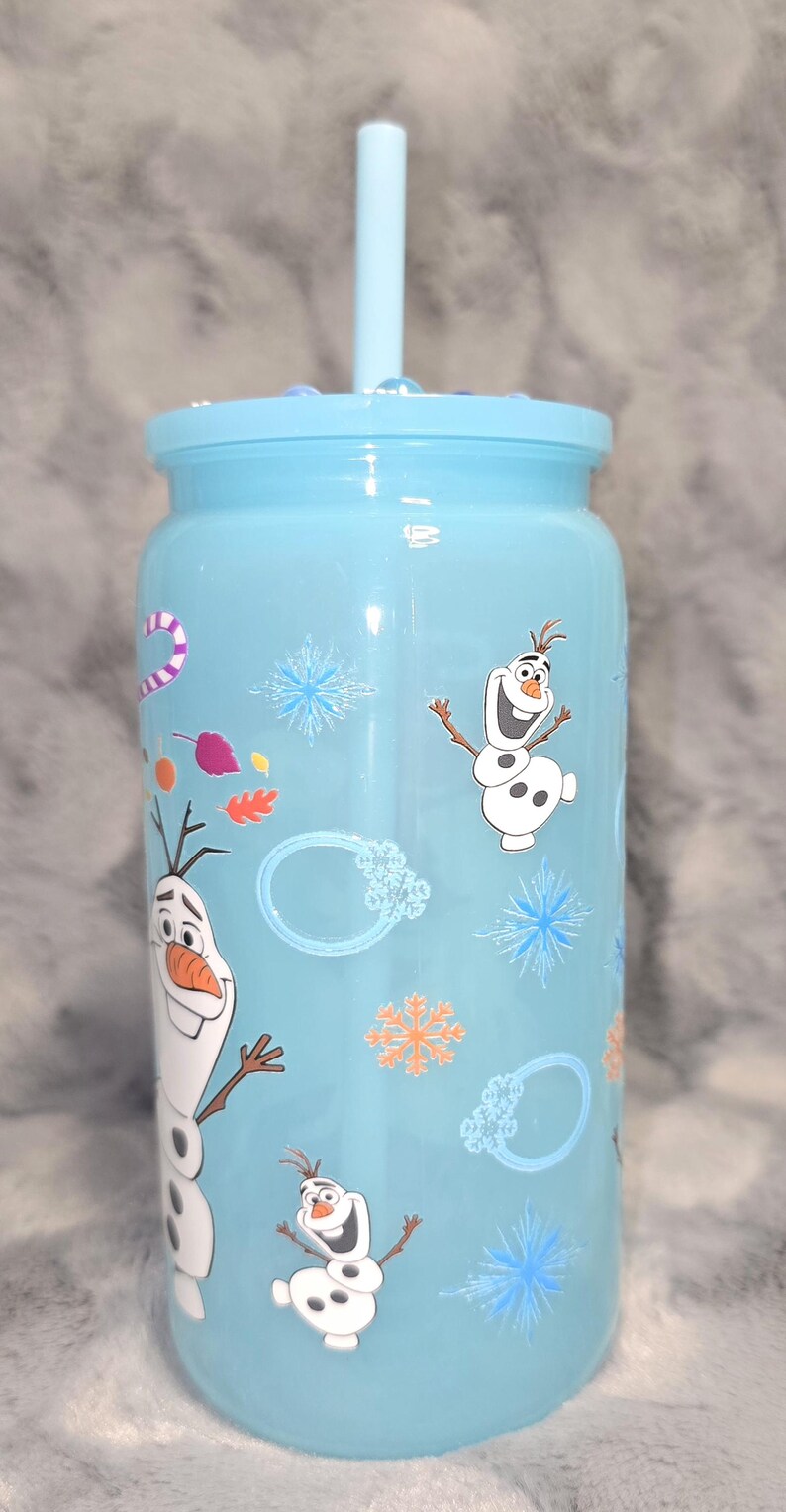 May include: A light blue tumbler with a matching lid and straw. The tumbler is decorated with images of Olaf from the movie Frozen, snowflakes, and other winter-themed graphics. A fun and functional drinkware item.