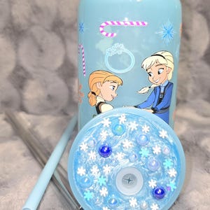 May include: A light blue tumbler with a lid and straw, featuring illustrations of two cartoon characters. The lid is decorated with snowflakes and blue beads. A candy cane and ring graphic are also on the tumbler.
