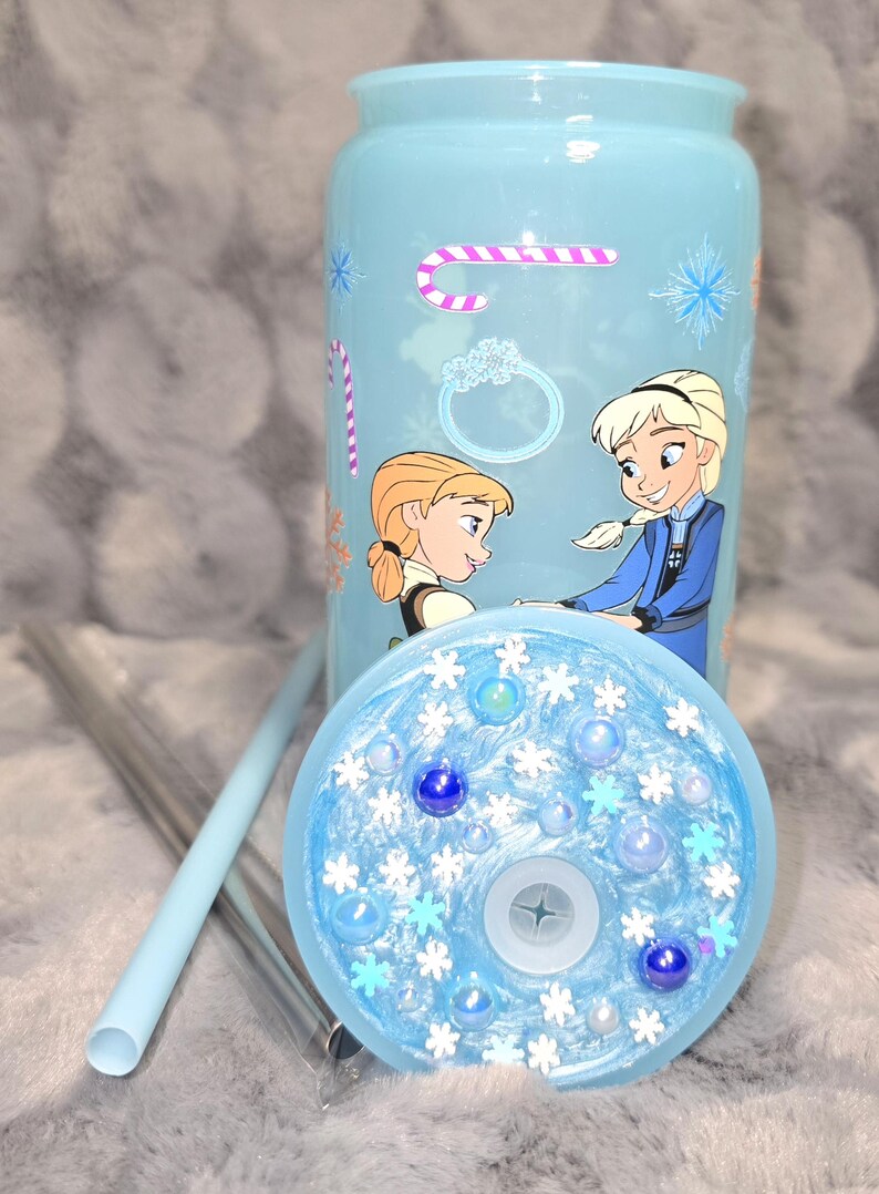 May include: A light blue tumbler with a lid and straw, featuring illustrations of two cartoon characters. The lid is decorated with snowflakes and blue beads. A candy cane and ring graphic are also on the tumbler.
