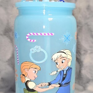 May include: A light blue tumbler with a lid and straw, decorated with cartoon images of two girls. The tumbler has a winter theme with snowflake and candy cane designs. The tumbler is approximately 7 inches tall.