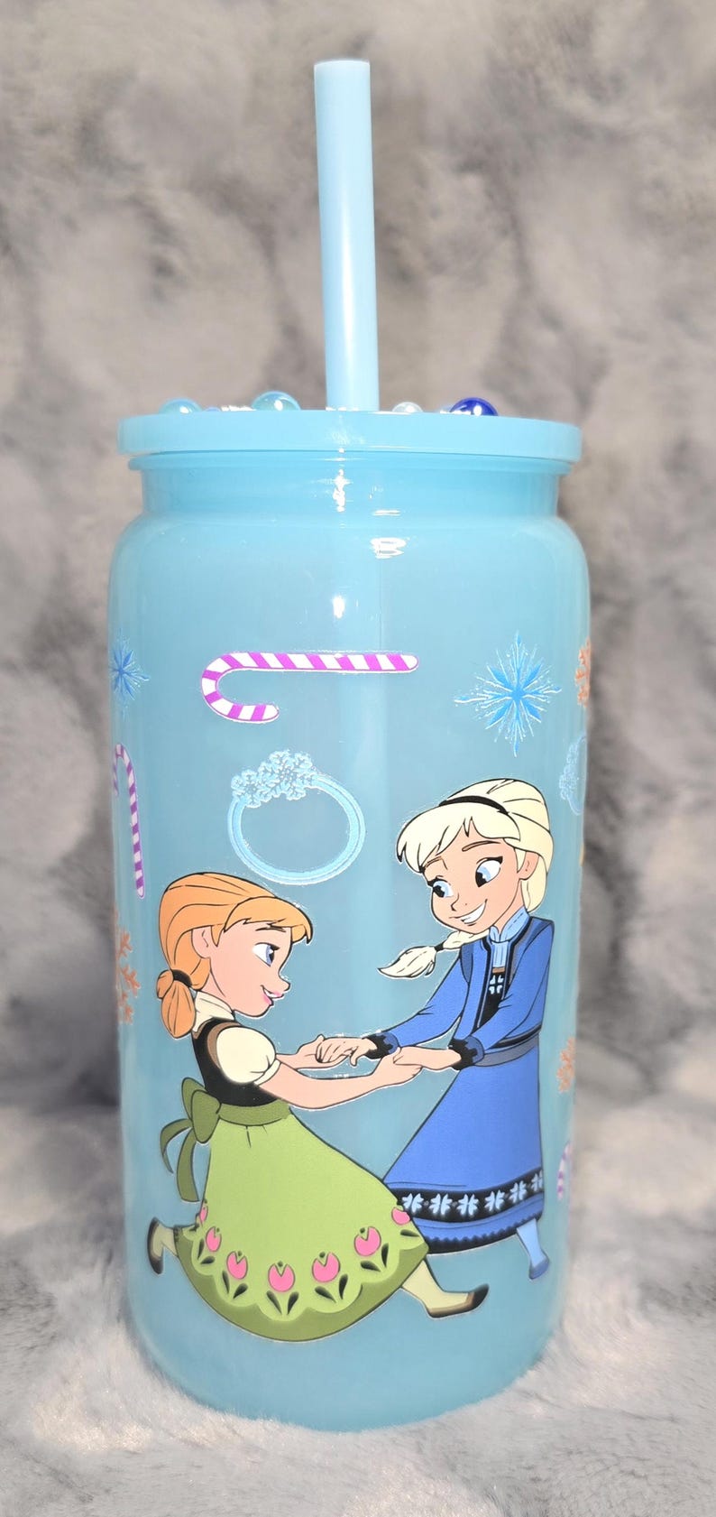 May include: A light blue tumbler with a lid and straw, decorated with cartoon images of two girls. The tumbler has a winter theme with snowflake and candy cane designs. The tumbler is approximately 7 inches tall.