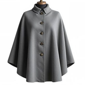 Op de afbeelding: Een grijze cape met een klassiek design. De cape heeft een kraag, een knoopsluiting aan de voorkant en een vloeiend silhouet. De knopen hebben een donkerdere bruine tint. De cape wordt op een mannequin gepresenteerd.